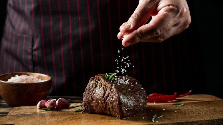 Hand som strör salt över en grillad köttbit på en träbräda